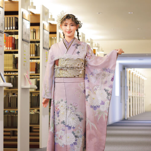 紅梅色の振袖に白菊と藤の花模様をあしらい、紫の帯締を合わせた女性の立ち姿