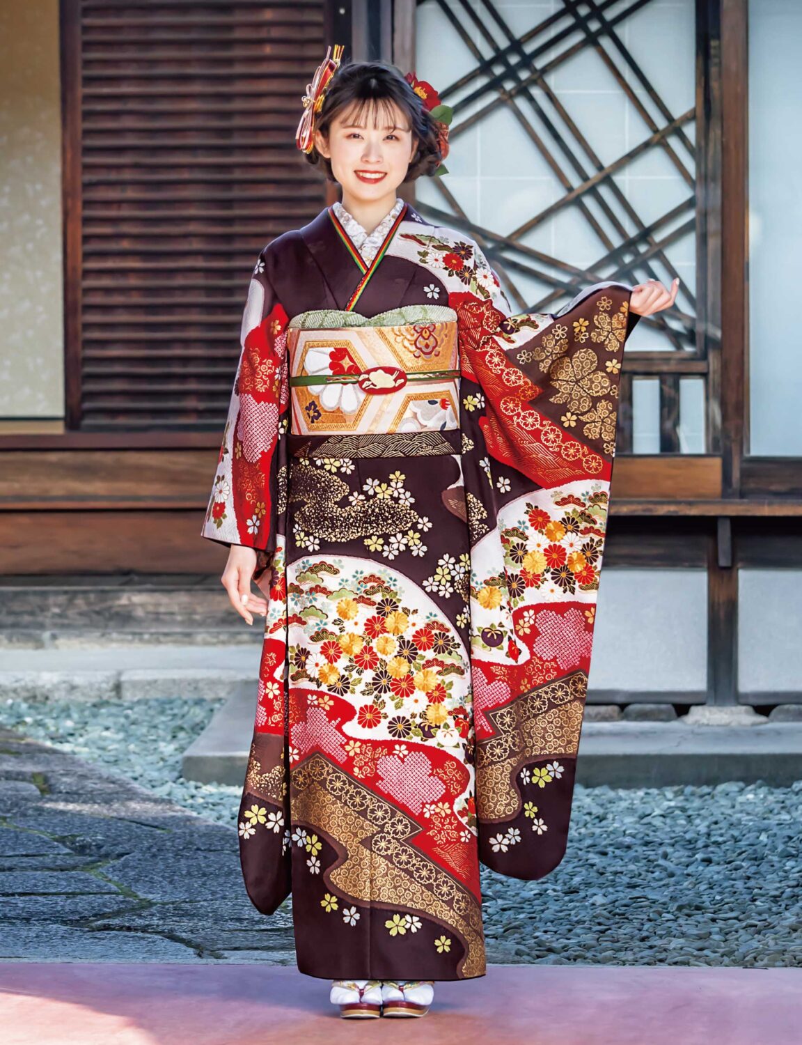 深紅と金彩の振袖を纏った女性が、花と幾何学模様の意匠と帯締の煌めきで華やかに装っている様子