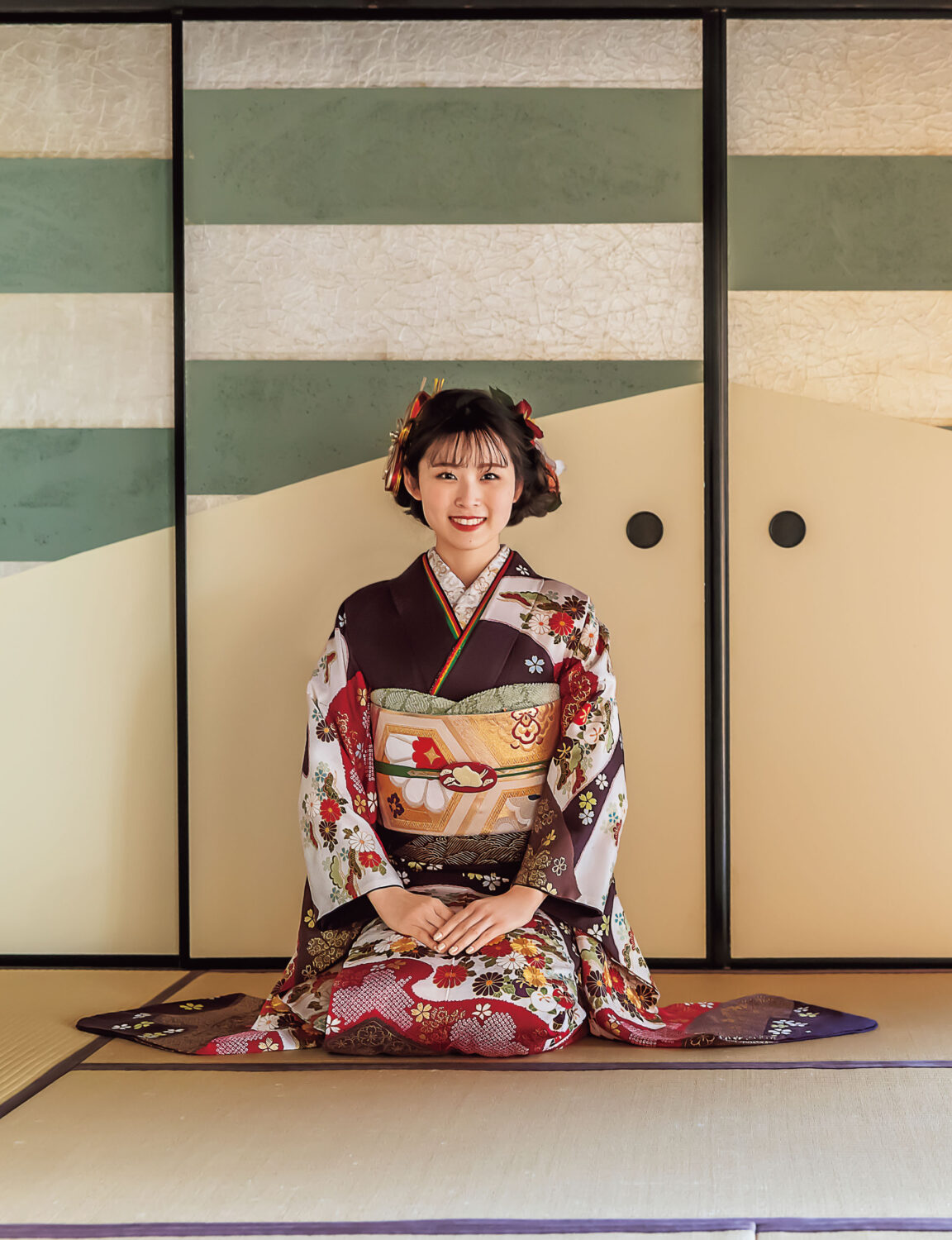 深紅と金彩の振袖を纏った女性が、花と幾何学模様の意匠と帯締の煌めきで華やかに装っている様子