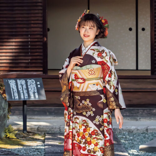 深紅と金彩の振袖を纏った女性が、花と幾何学模様の意匠と帯締の煌めきで華やかに装っている様子