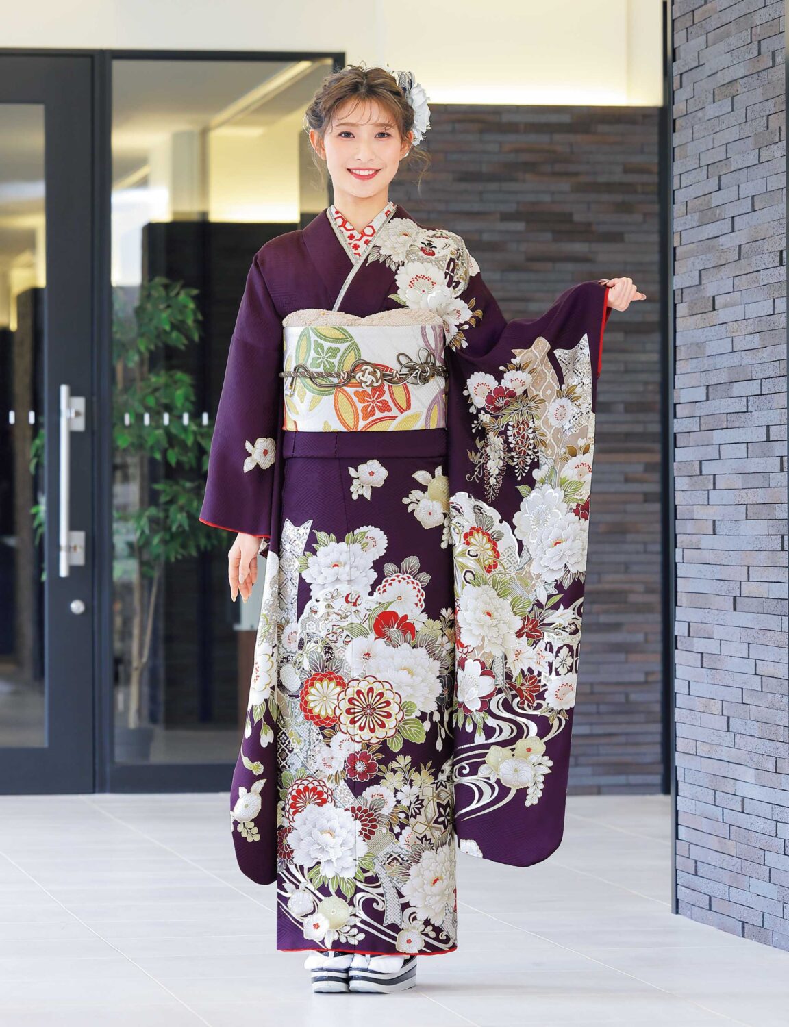 紫地に菊や牡丹が描かれた振袖姿の女性。白地に橙と緑の帯、彩り豊かな帯締が華やかさを添える。