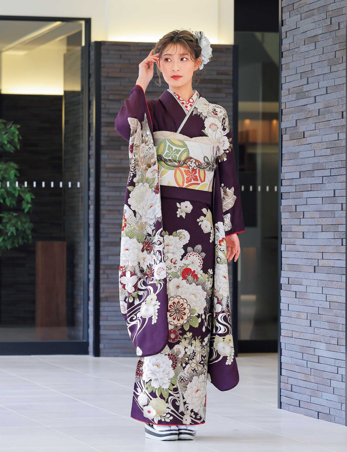 紫地に菊や牡丹が描かれた振袖姿の女性。白地に橙と緑の帯、彩り豊かな帯締が華やかさを添える。