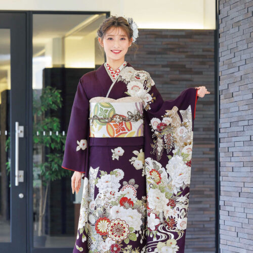 紫地に菊や牡丹が描かれた振袖姿の女性。白地に橙と緑の帯、彩り豊かな帯締が華やかさを添える。