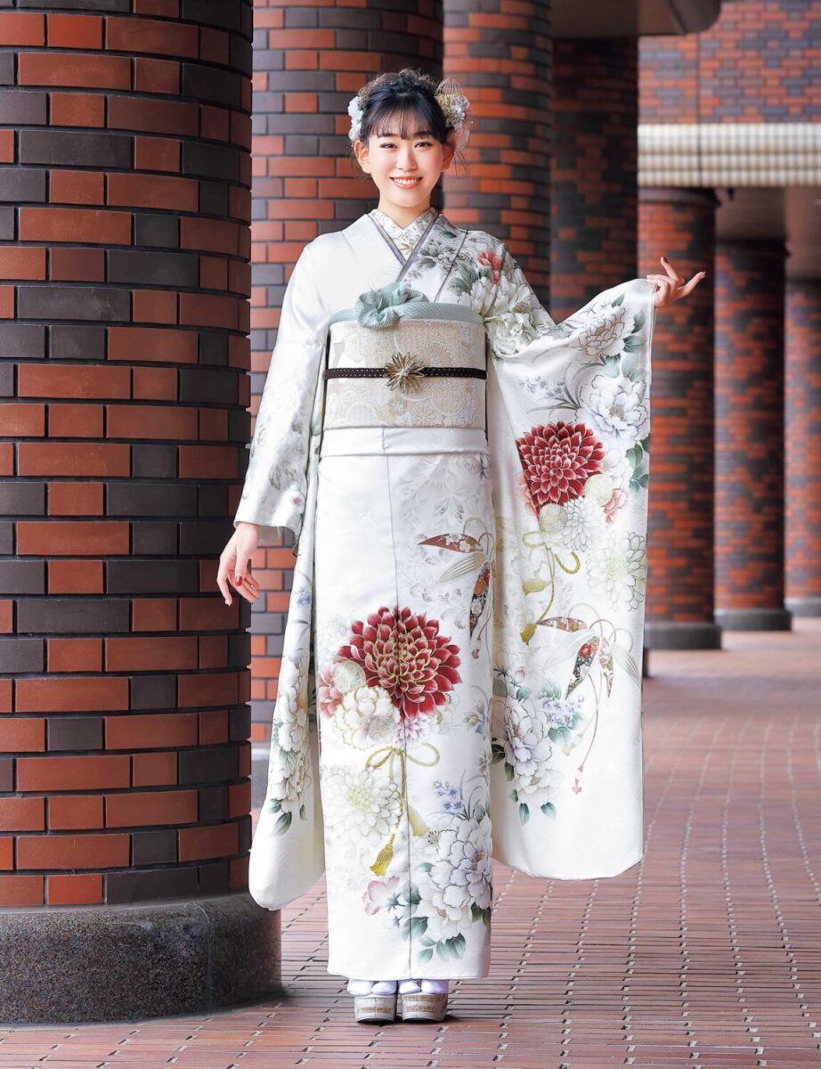 白地に赤・緑・金の菊模様が施された振袖姿の女性。帯締がアクセントになった華やかな装い。