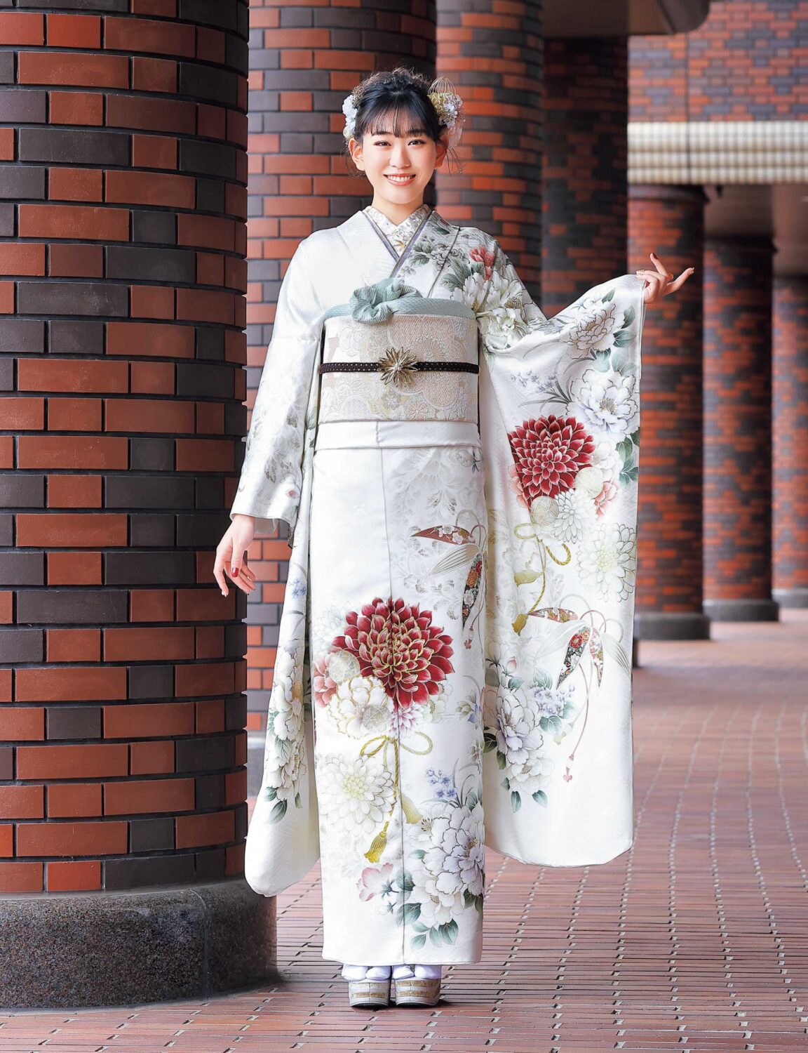 白地に赤・緑・金の菊模様が施された振袖姿の女性。帯締がアクセントになった華やかな装い。