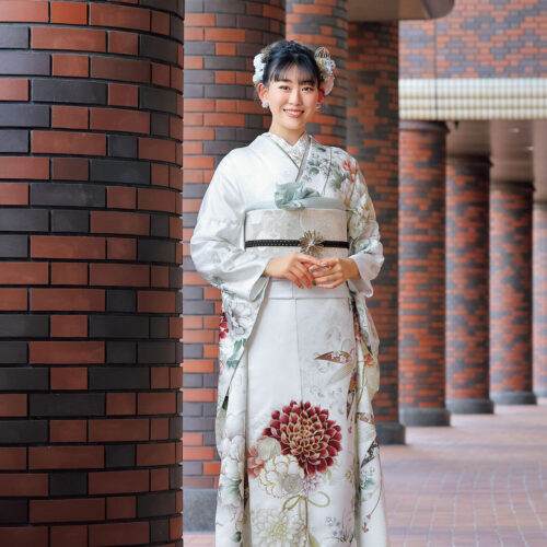 白地に赤・緑・金の菊模様が施された振袖姿の女性。帯締がアクセントになった華やかな装い。