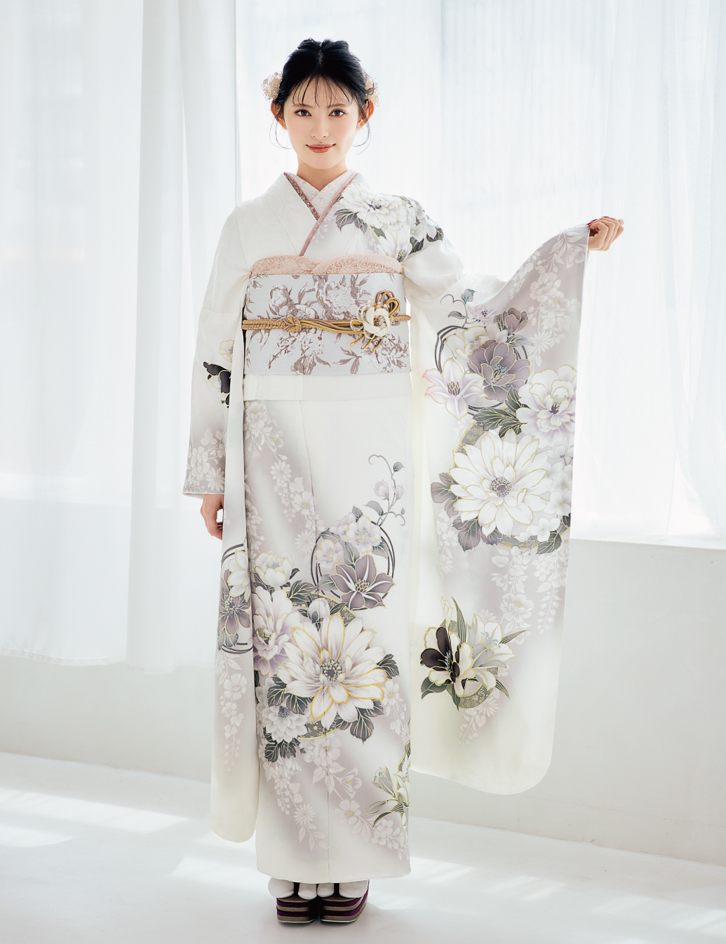 白地に灰色と淡紫の花柄をあしらい、淡色の帯と金の帯締を合わせた女性が、上品な振袖姿を見せている様子。