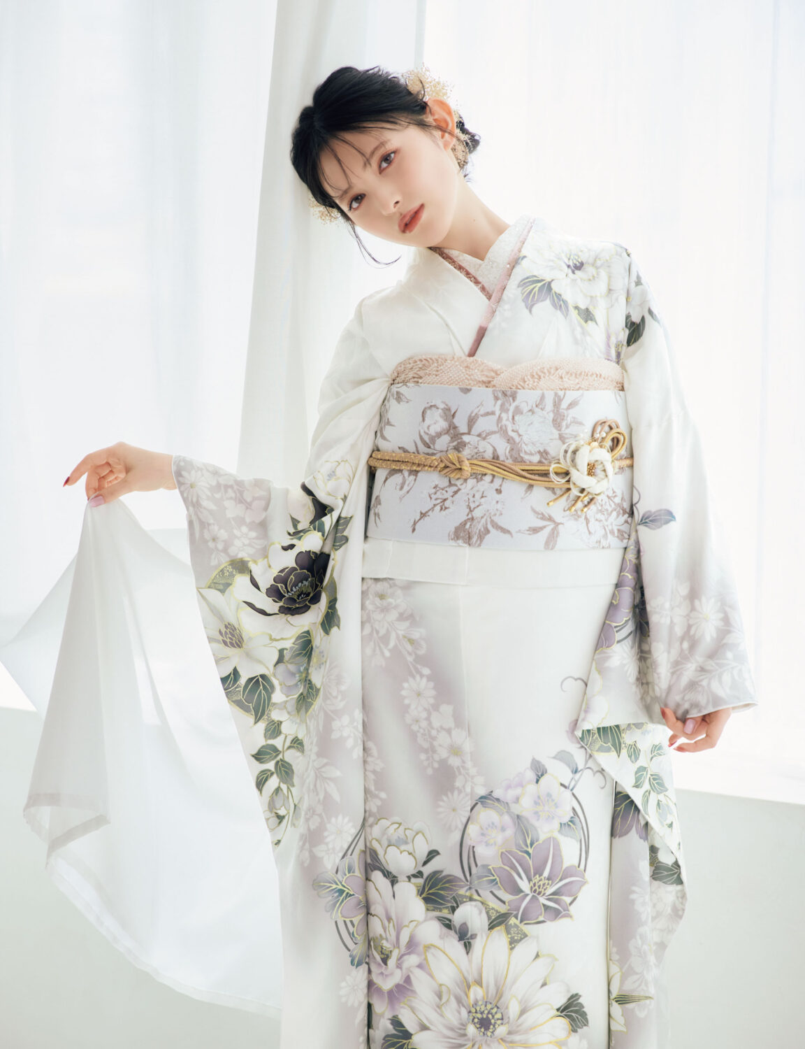 白地に灰色と淡紫の花柄をあしらい、淡色の帯と金の帯締を合わせた女性が、上品な振袖姿を見せている様子。