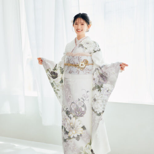 白地に灰色と淡紫の花柄をあしらい、淡色の帯と金の帯締を合わせた女性が、上品な振袖姿を見せている様子。