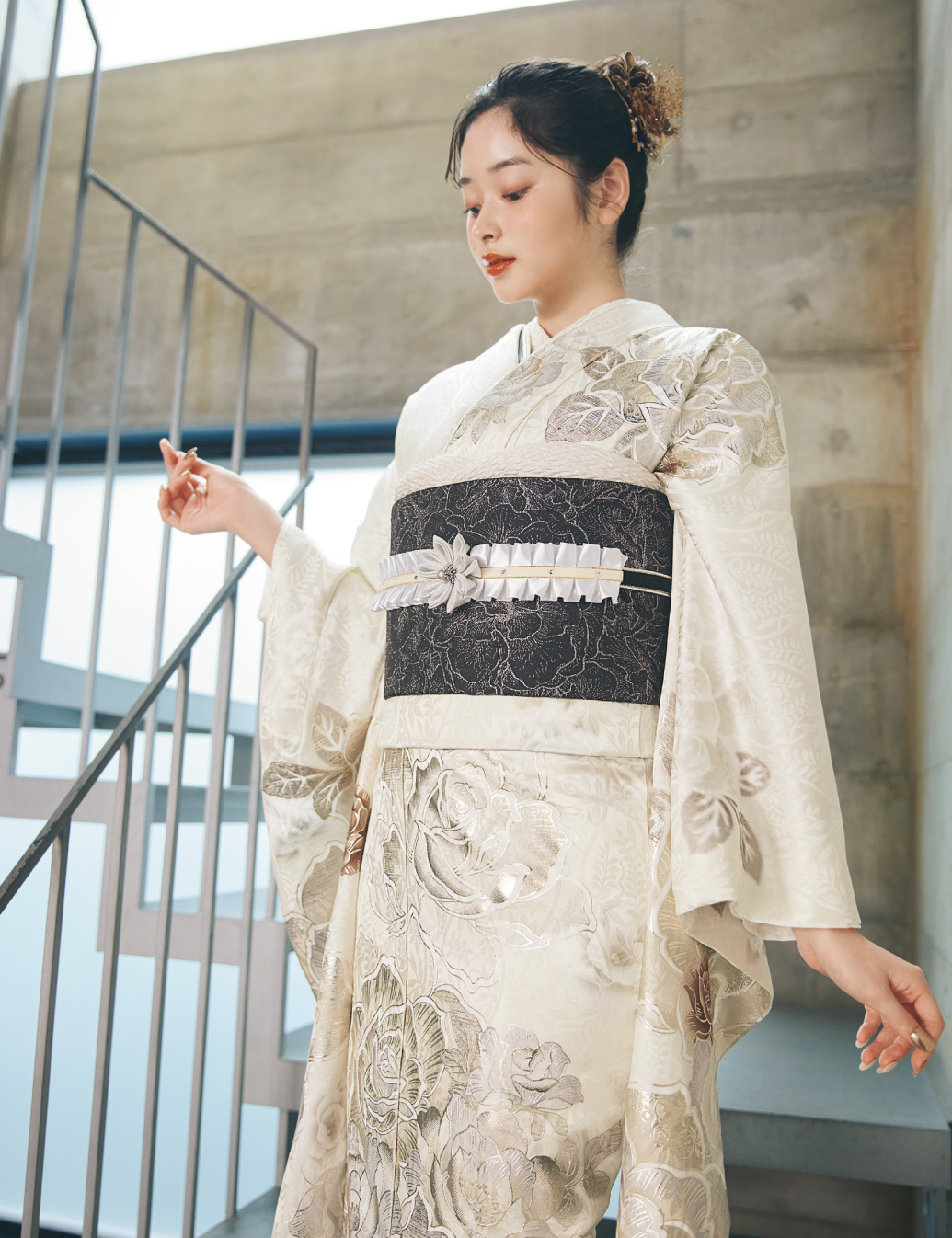 淡いクリーム色の振袖に金・銀・ベージュの花柄をあしらい、黒の帯と白の帯締を合わせた女性が、上品な振袖姿を見せている様子。