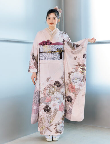 淡桃色の振袖に白・金・茶・モーブの花柄をあしらい、淡灰の帯と紫の帯締を合わせた女性が、優美で上品な振袖姿を見せている様子。