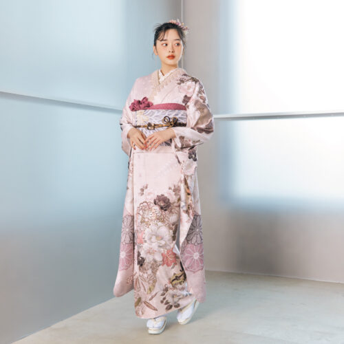 淡桃色の振袖に白・金・茶・モーブの花柄をあしらい、淡灰の帯と紫の帯締を合わせた女性が、優美で上品な振袖姿を見せている様子。