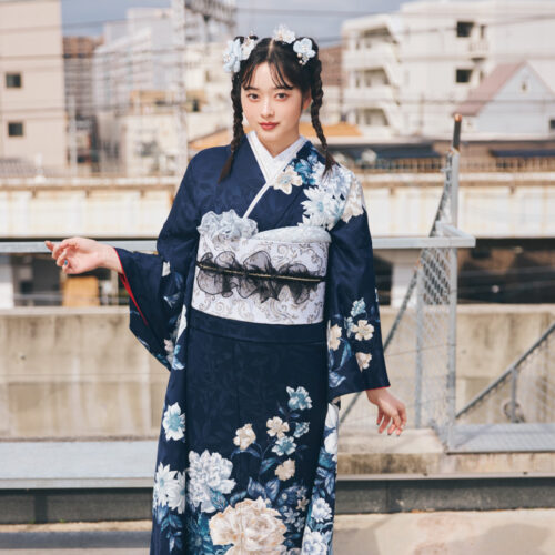 深紺の振袖に白・水色・ベージュの花柄をあしらい、白帯と黒リボンの帯締を合わせた女性が、上品で都会的な振袖姿を見せている様子。