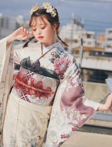 淡いクリーム色の振袖にピンク・バーガンディ・グレーの花柄をあしらい、重ね帯と黒レース調の帯締を合わせた女性が、上品で華やかな振袖姿を見せている様子。