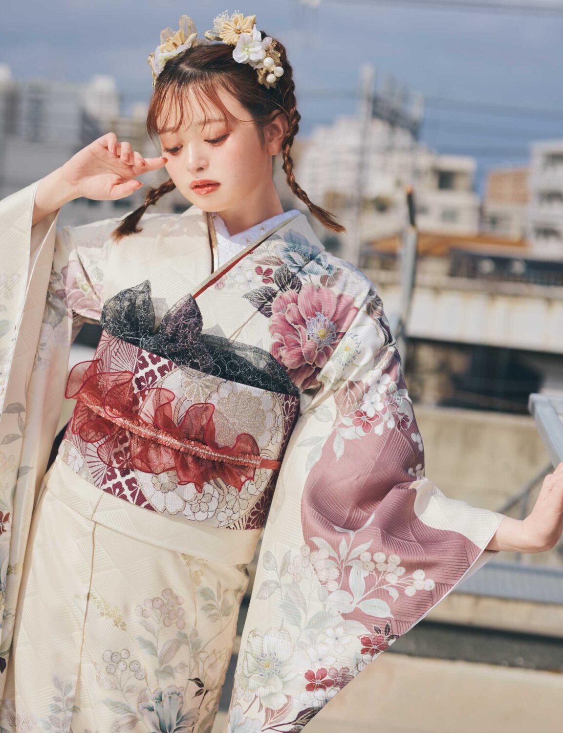 淡いクリーム色の振袖にピンク・バーガンディ・グレーの花柄をあしらい、重ね帯と黒レース調の帯締を合わせた女性が、上品で華やかな振袖姿を見せている様子。