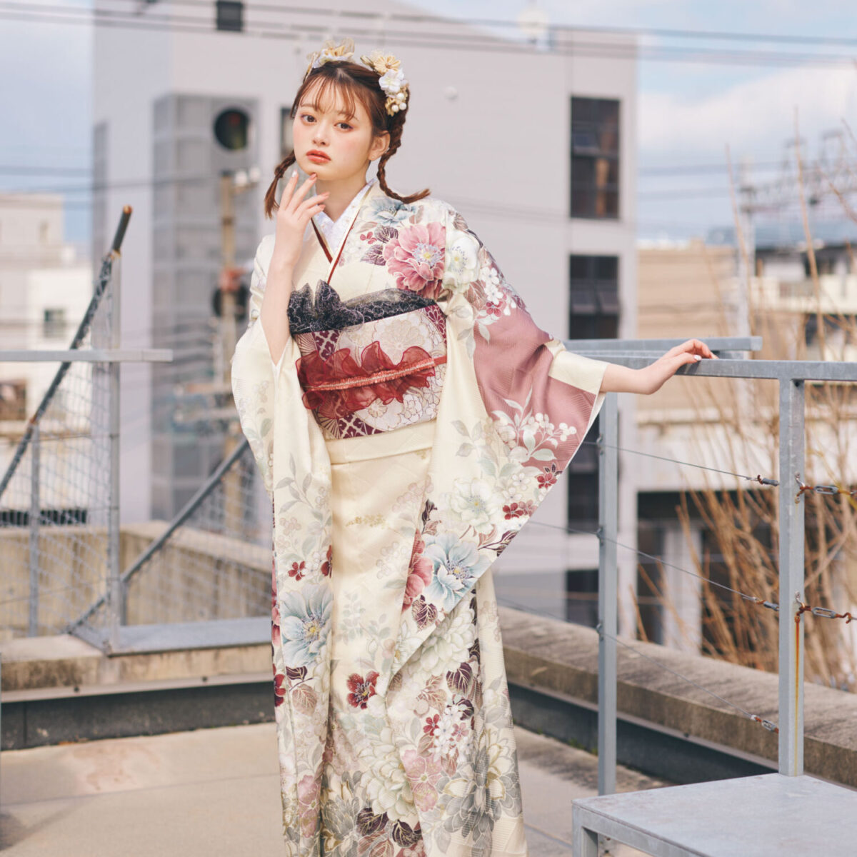 淡いクリーム色の振袖にピンク・バーガンディ・グレーの花柄をあしらい、重ね帯と黒レース調の帯締を合わせた女性が、上品で華やかな振袖姿を見せている様子。
