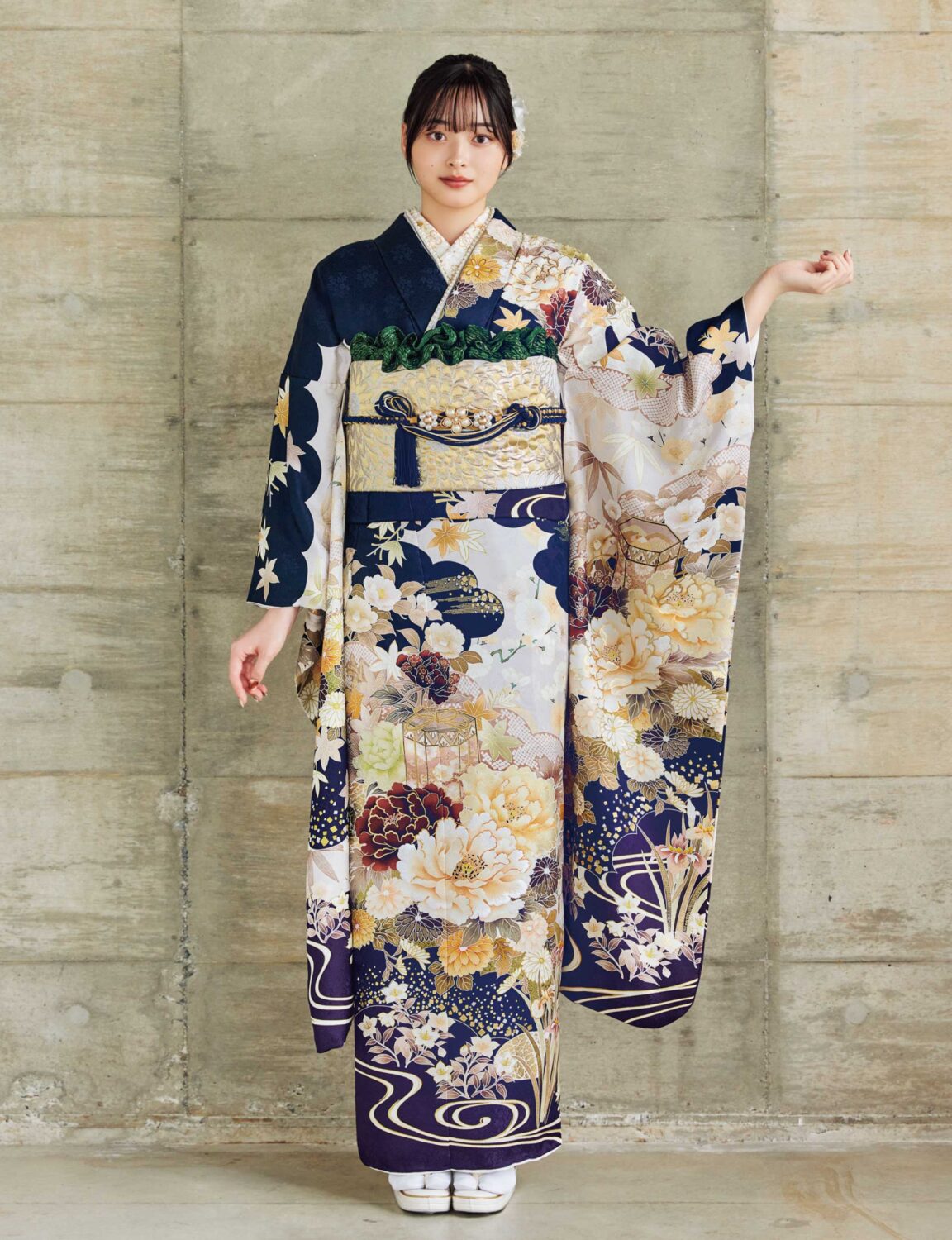金・クリーム・白・紺・緑の花柄をあしらった振袖に、淡金の帯と青の帯締を合わせた女性が、重厚で上品な振袖姿を見せている様子。