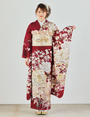 深紅の振袖に白・生成り・金の花柄をあしらい、淡金の帯と装飾的な帯飾りを合わせた女性が、華やかで上質な振袖姿を見せている様子。
