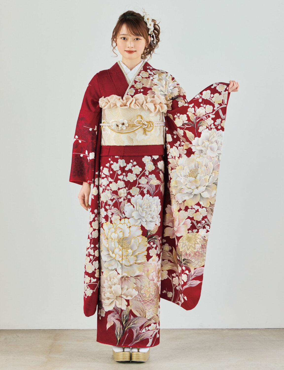 深紅の振袖に白・生成り・金の花柄をあしらい、淡金の帯と装飾的な帯飾りを合わせた女性が、華やかで上質な振袖姿を見せている様子。