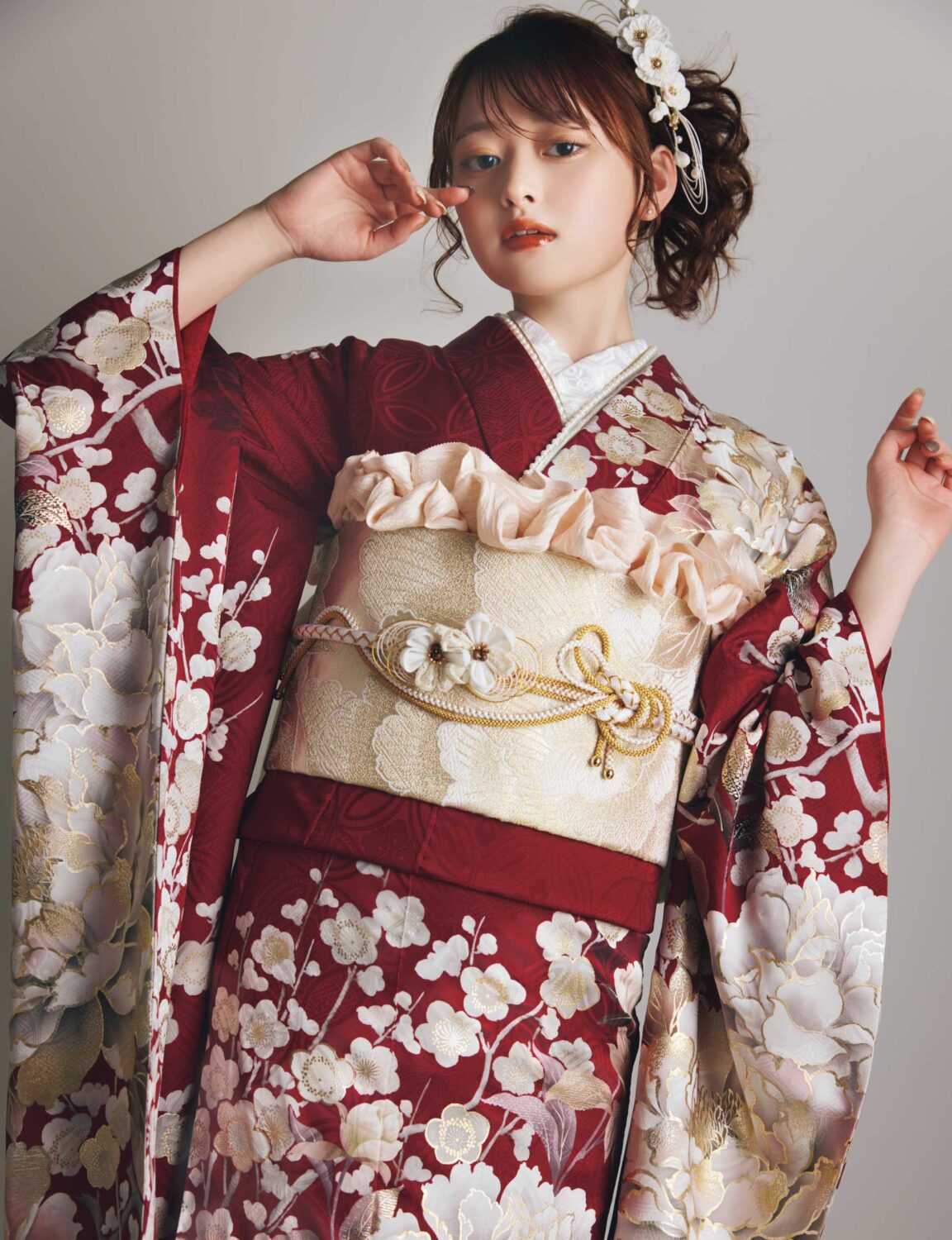 深紅の振袖に白・生成り・金の花柄をあしらい、淡金の帯と装飾的な帯飾りを合わせた女性が、華やかで上質な振袖姿を見せている様子。