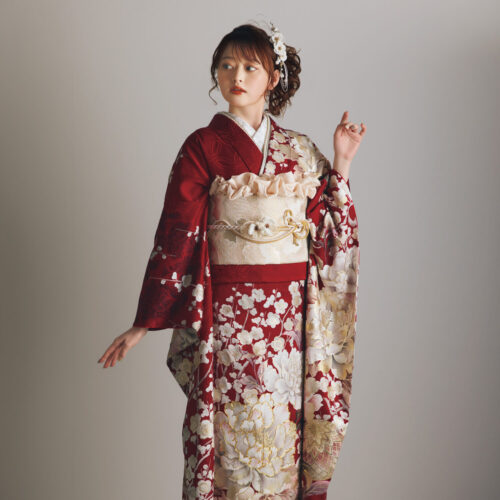 深紅の振袖に白・生成り・金の花柄をあしらい、淡金の帯と装飾的な帯飾りを合わせた女性が、華やかで上質な振袖姿を見せている様子。