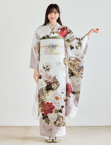 白地の振袖に赤・金・緑・ベージュの花柄をあしらい、淡色の帯と花の帯飾りを合わせた女性が、上品で華やかな振袖姿を見せている様子。