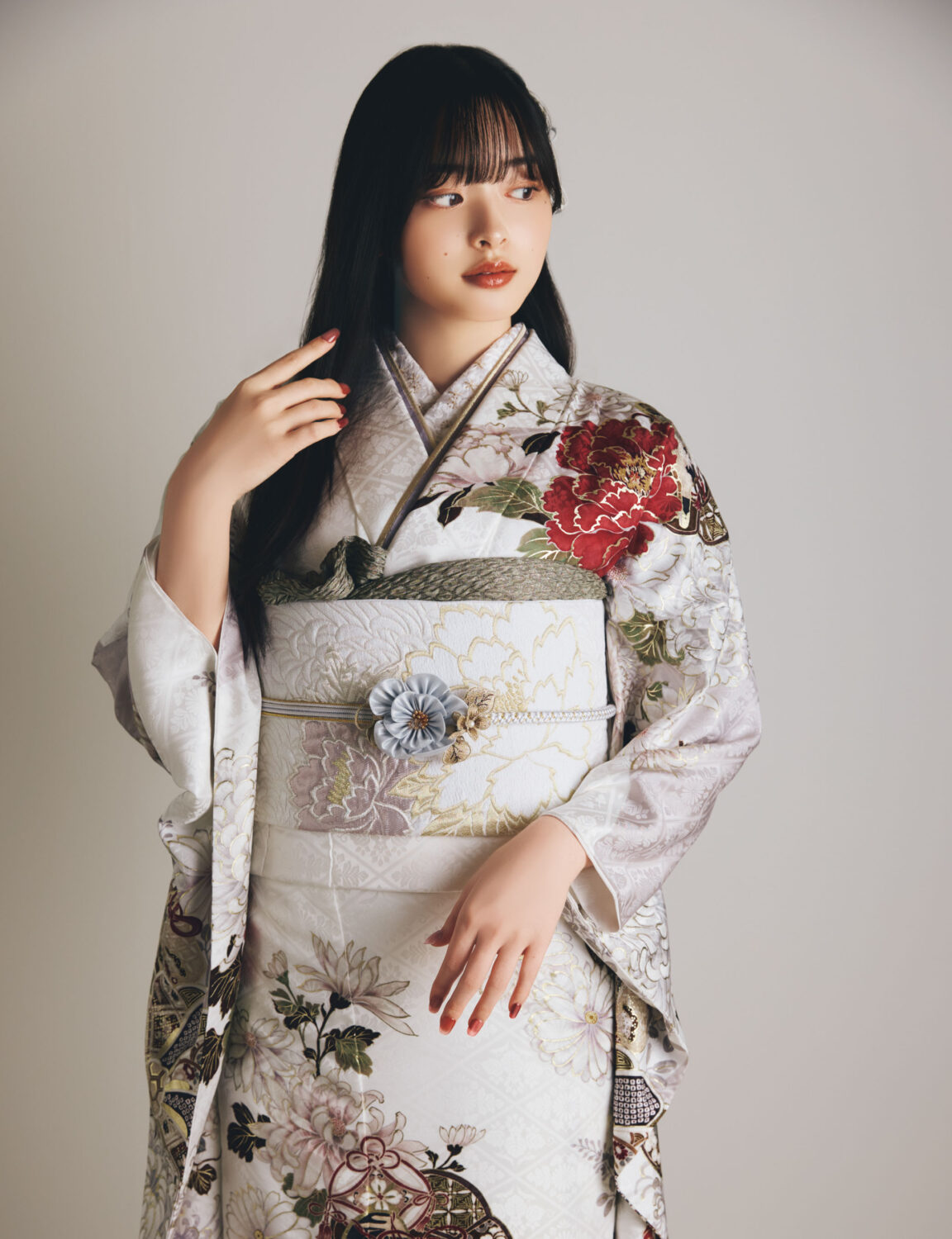 白地の振袖に赤・金・緑・ベージュの花柄をあしらい、淡色の帯と花の帯飾りを合わせた女性が、上品で華やかな振袖姿を見せている様子。