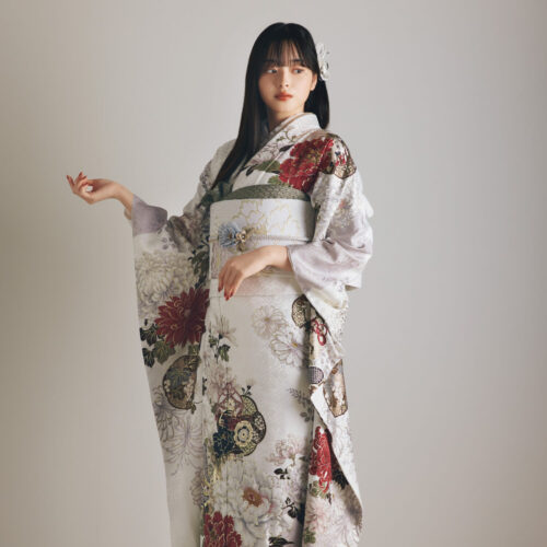白地の振袖に赤・金・緑・ベージュの花柄をあしらい、淡色の帯と花の帯飾りを合わせた女性が、上品で華やかな振袖姿を見せている様子。