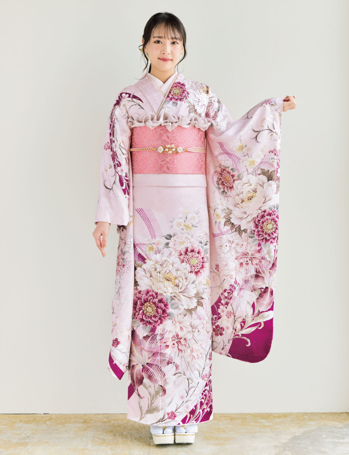 淡桃色の振袖に白・生成り・深紅の花柄をあしらい、ピンク系の帯と帯飾りを合わせた女性が、優美で可憐な振袖姿を見せている様子。