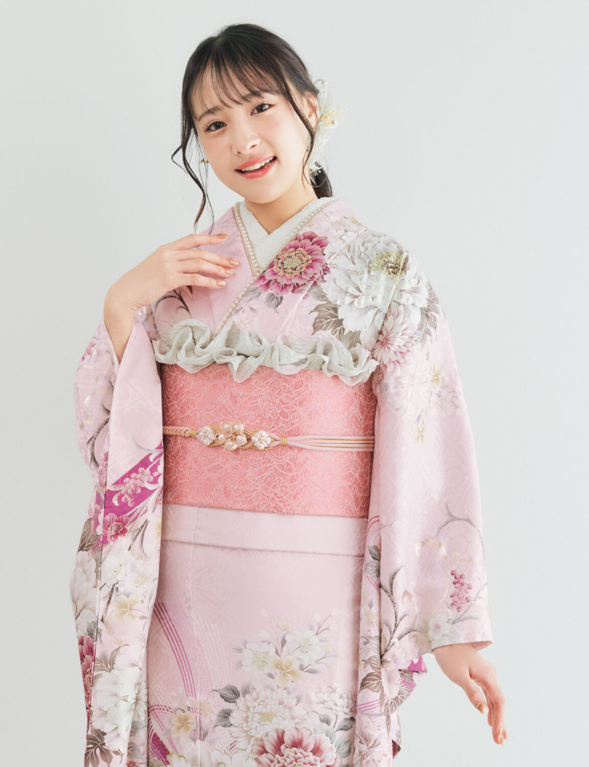 淡桃色の振袖に白・生成り・深紅の花柄をあしらい、ピンク系の帯と帯飾りを合わせた女性が、優美で可憐な振袖姿を見せている様子。