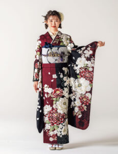 えんじと黒の振袖に白紅の牡丹と幾何文様をあしらい、青の帯締を合わせた女性の立ち姿