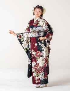 えんじと黒の振袖に白紅の牡丹と幾何文様をあしらい、青の帯締を合わせた女性の立ち姿