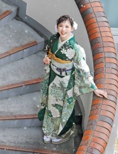 深緑の振袖に白牡丹と菊の花模様をあしらい、金の帯と濃紺の帯締を合わせた女性の立ち姿