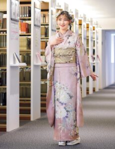 紅梅色の振袖に白菊と藤の花模様をあしらい、紫の帯締を合わせた女性の立ち姿
