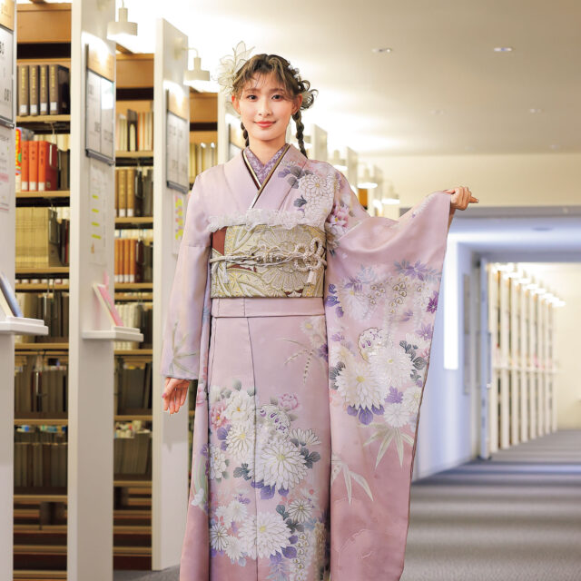 紅梅色の振袖に白菊と藤の花模様をあしらい、紫の帯締を合わせた女性の立ち姿