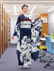 銀白の花模様が描かれた深藍の振袖を着た女性が図書館風の空間に立つ姿。赤の帯締がアクセント。