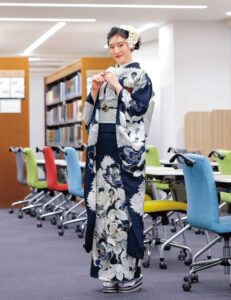 銀白の花模様が描かれた深藍の振袖を着た女性が図書館風の空間に立つ姿。赤の帯締がアクセント。