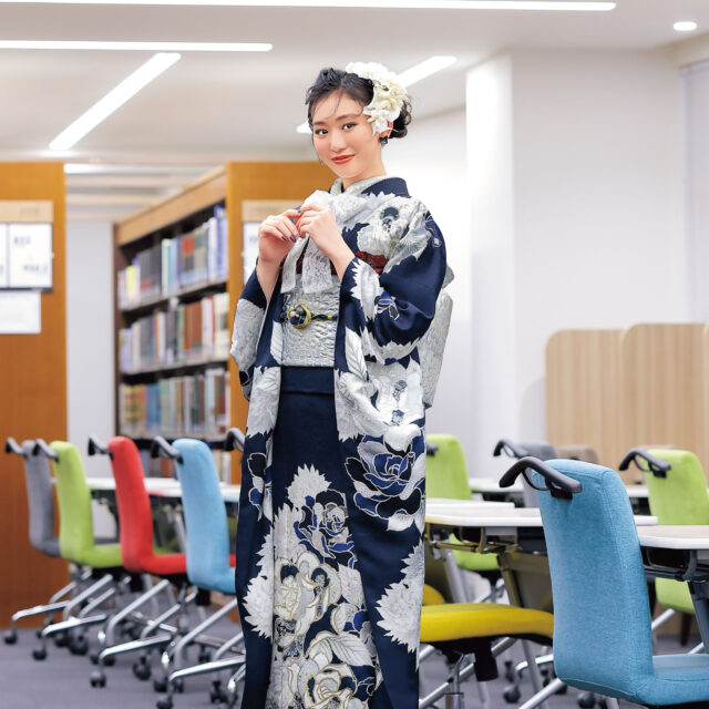 銀白の花模様が描かれた深藍の振袖を着た女性が図書館風の空間に立つ姿。赤の帯締がアクセント。