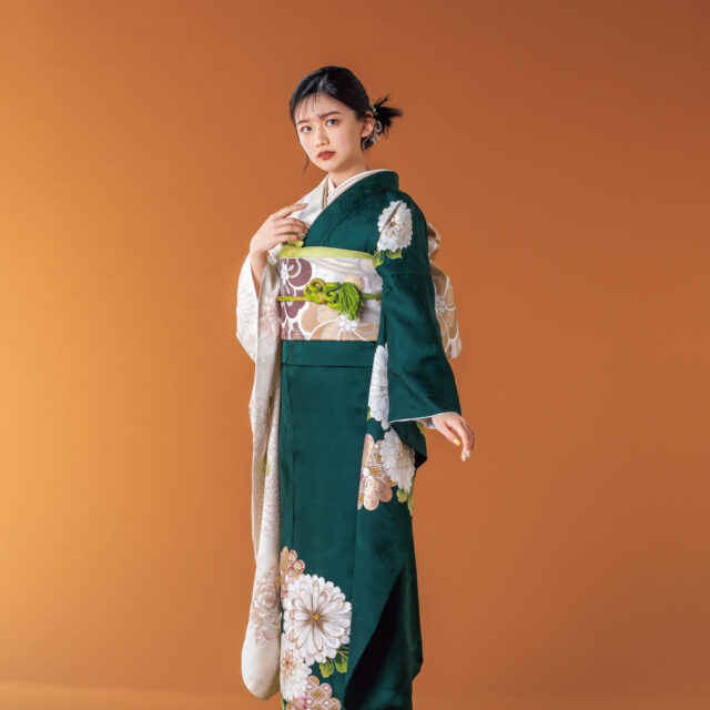 深緑と白の振袖に菊とダリアをあしらい、淡彩の帯と帯締を合わせた女性の立ち姿